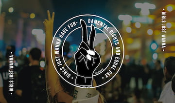 The sticker of the Girls Just Wanna Have Fun-damental Rights and Equal Pay movement on top of the picture of a woman showing the peace sign during a protest.